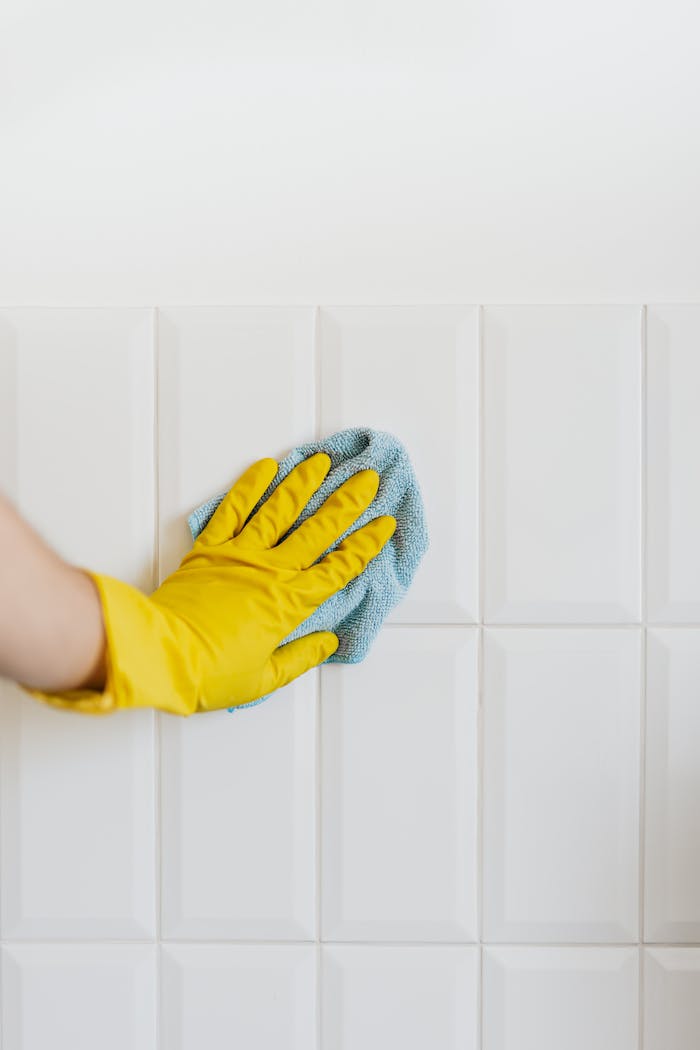 Crop unrecognizable person in yellow rubber gloves cleaning white tile wall with blue microfiber cloth in daylight