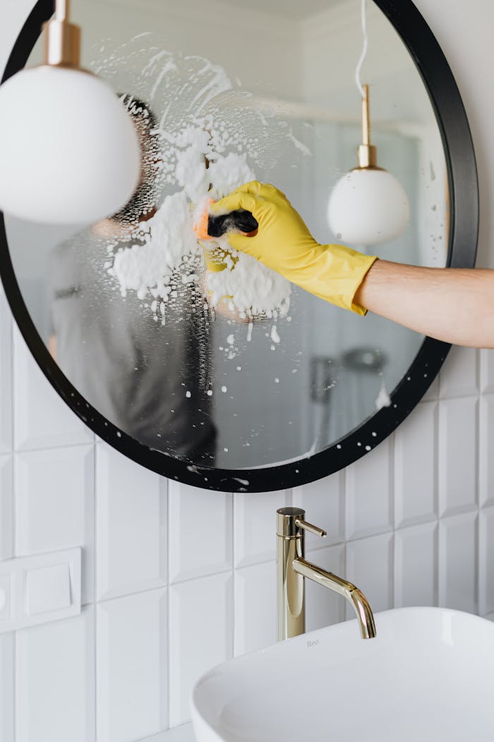 Anonymous housekeeper in yellow rubber gloves wiping round mirror by sponge in bathroom
